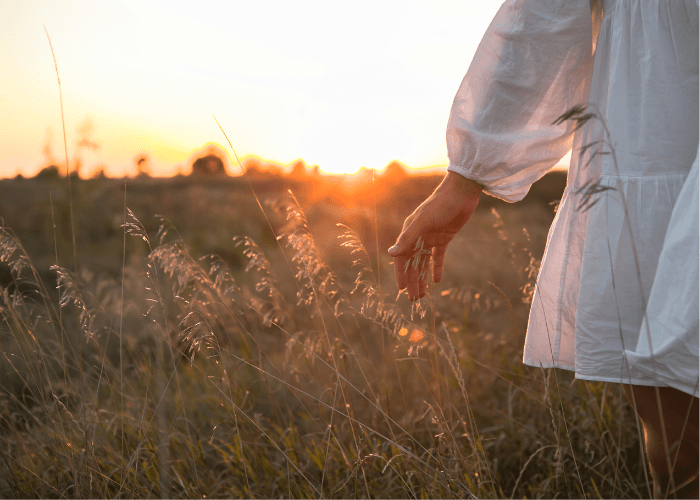 Eine junge Frau im weißen Kleid geht im Abendrot auf einem Feld spazieren und denkt über ihre Partnerschaft nach